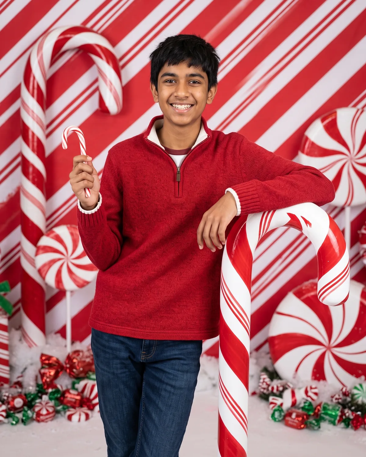 Studio photo of the same smiling teenage boy with short black hair in a red quarter-zip knit sweater and blue jeans, holding a small candy cane and leaning on a giant red-and-white candy cane, in front of a diagonal peppermint-striped backdrop with giant swirl lollipops and wrapped candies on fluffy white fake snow.