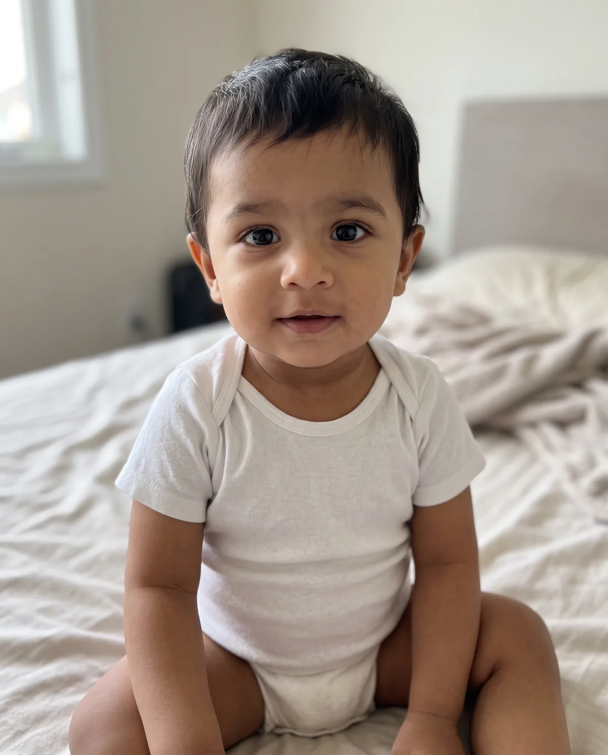 South Asian baby boy with short dark hair in a white onesie, sitting on a bed with beige sheets.