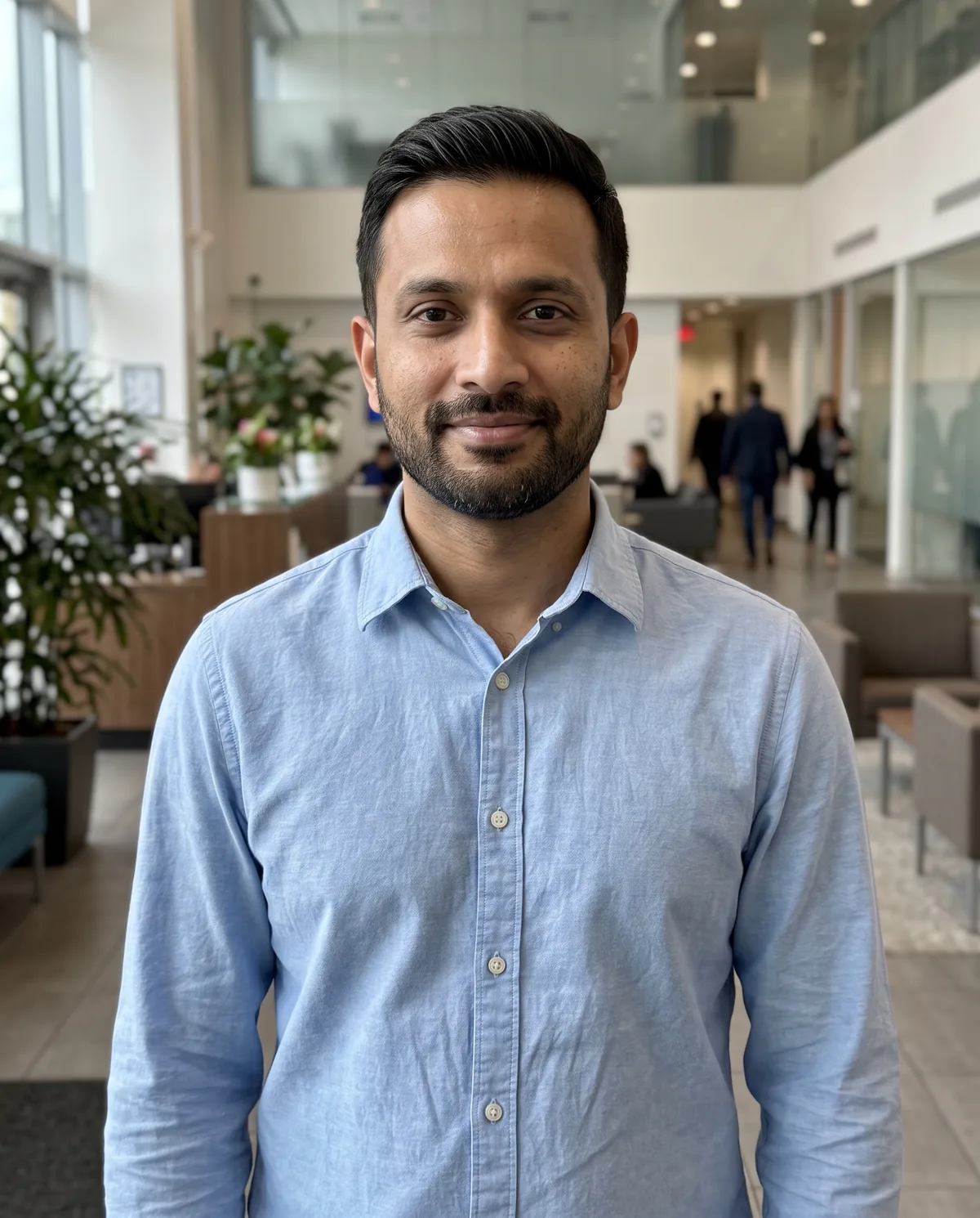South Asian man with short dark hair and a trimmed beard in a light-blue button shirt, standing in a modern office lobby.