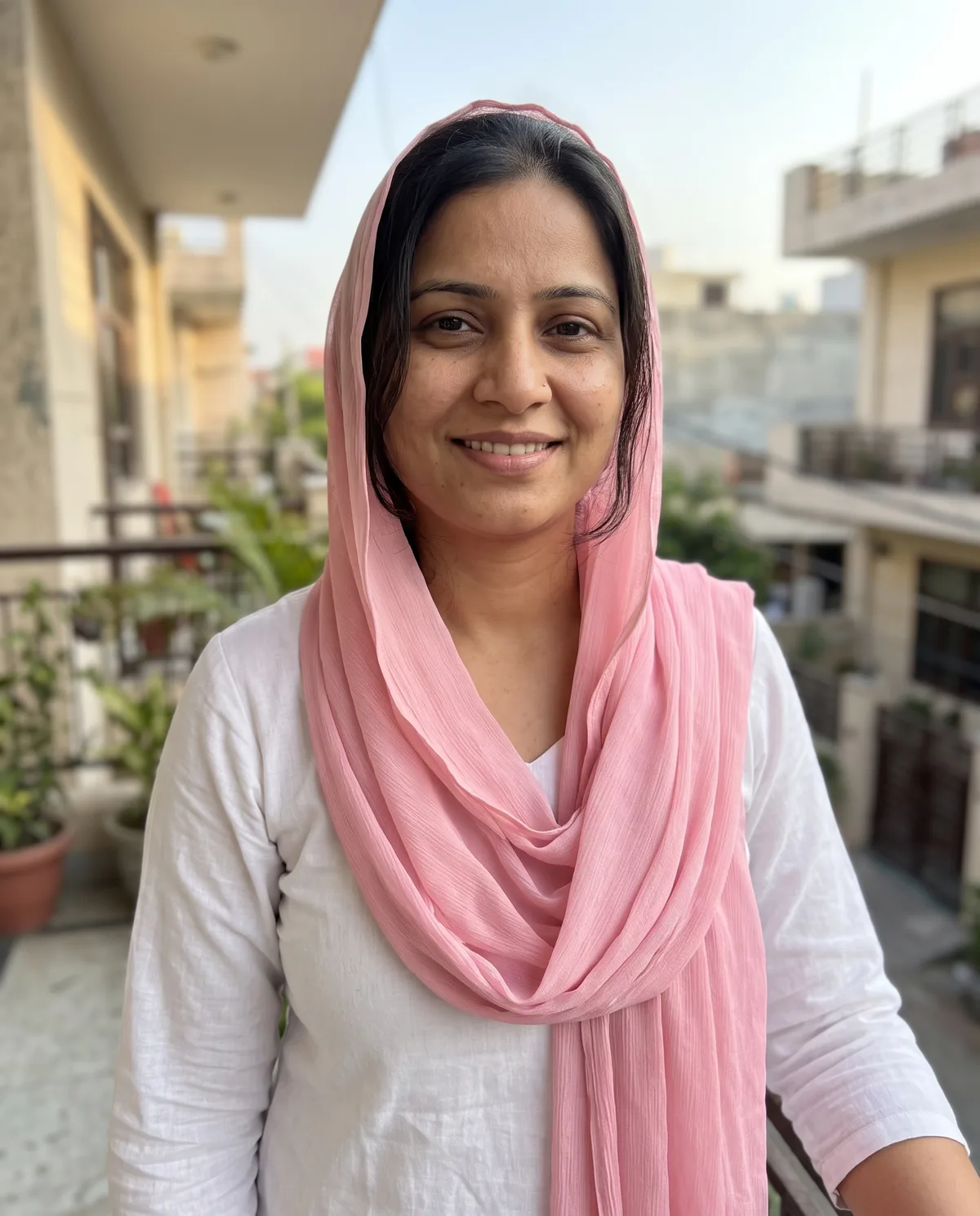 South Asian woman in a soft-pink dupatta over her hair and a white kurti, smiling on a home balcony.
