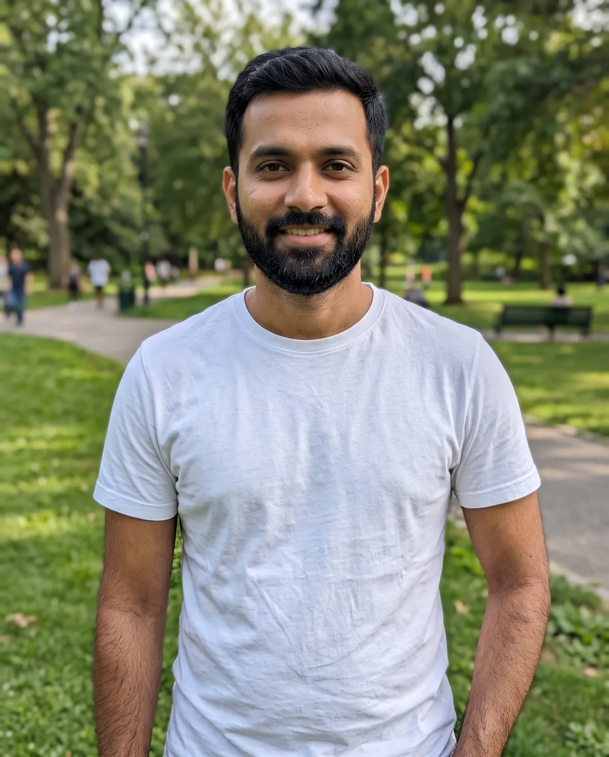 South Asian man with short dark hair and a full black beard in a white t-shirt, smiling in a green park.