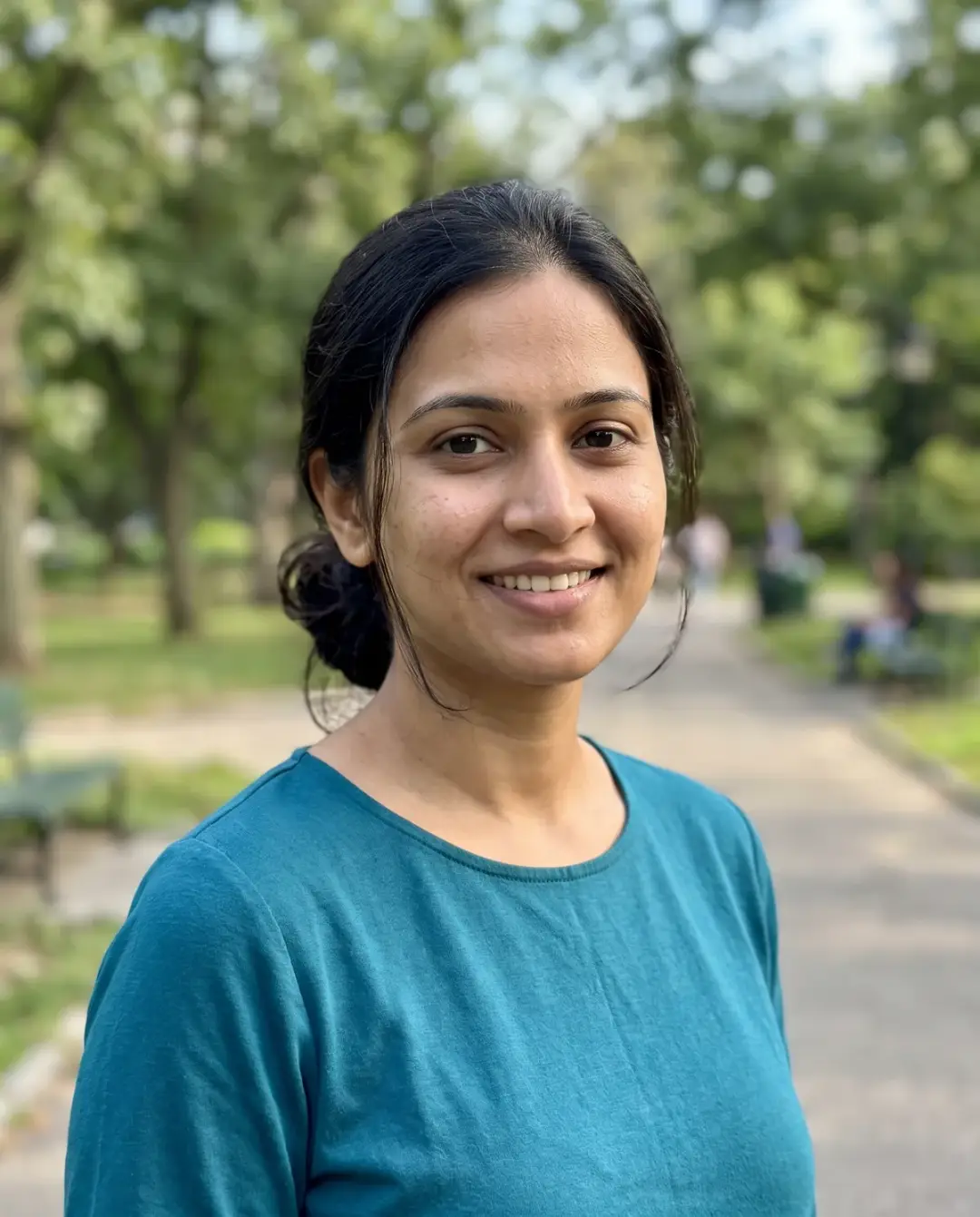 South Asian woman with dark hair in a low bun in a teal top, smiling in a tree-lined park.