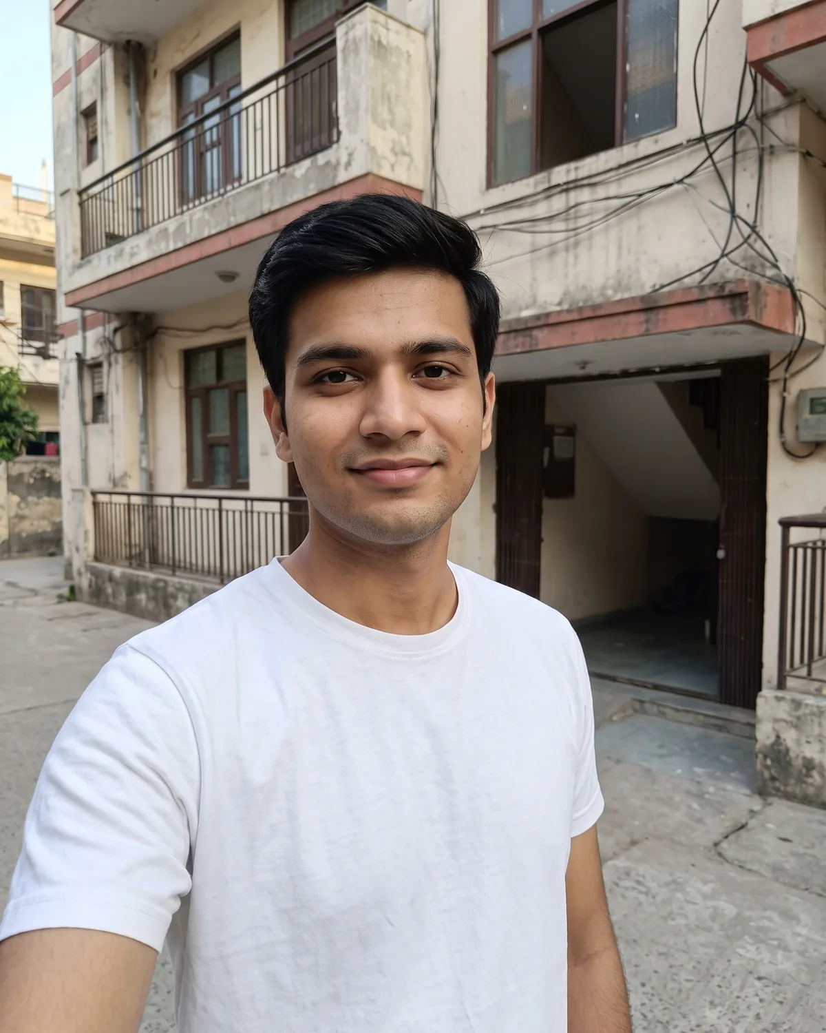 Young South Asian man with short side-parted black hair, clean shaven, in a plain white t-shirt, taking a selfie outside an older apartment building with balconies and overhead wires.