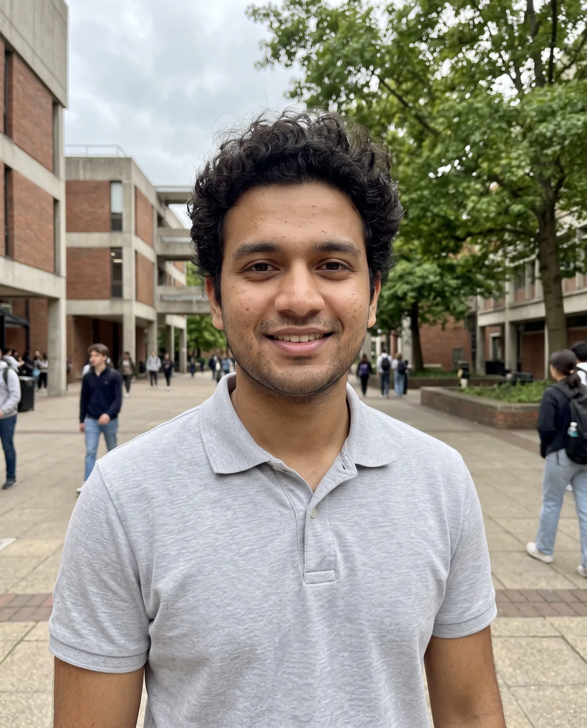 Young South Asian man with curly black hair and light stubble in a grey polo shirt, smiling in a brick university courtyard with students walking behind.