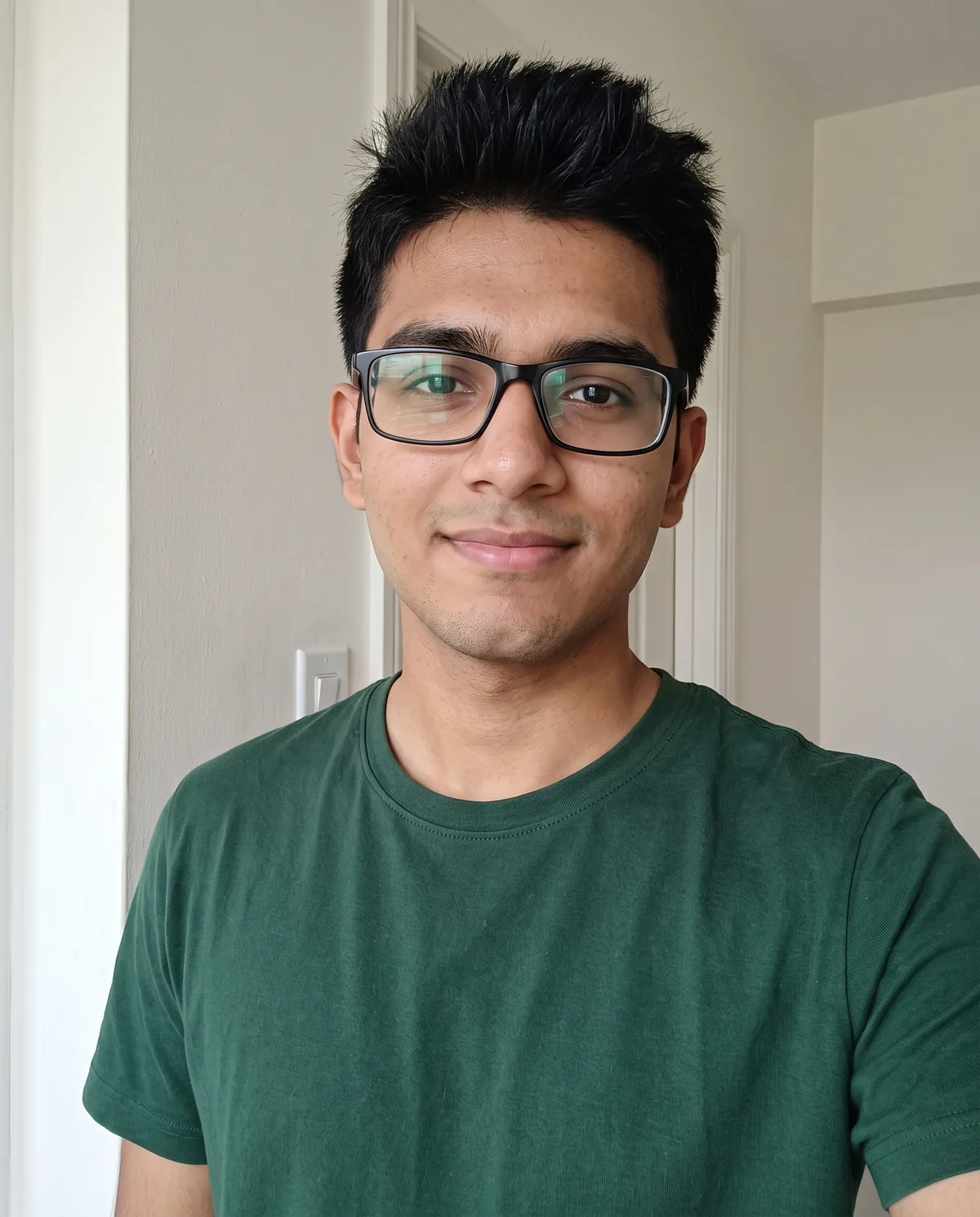 Young South Asian man with spiky black hair and rectangular black glasses in a dark green t-shirt, slight smile, indoors by a plain wall.
