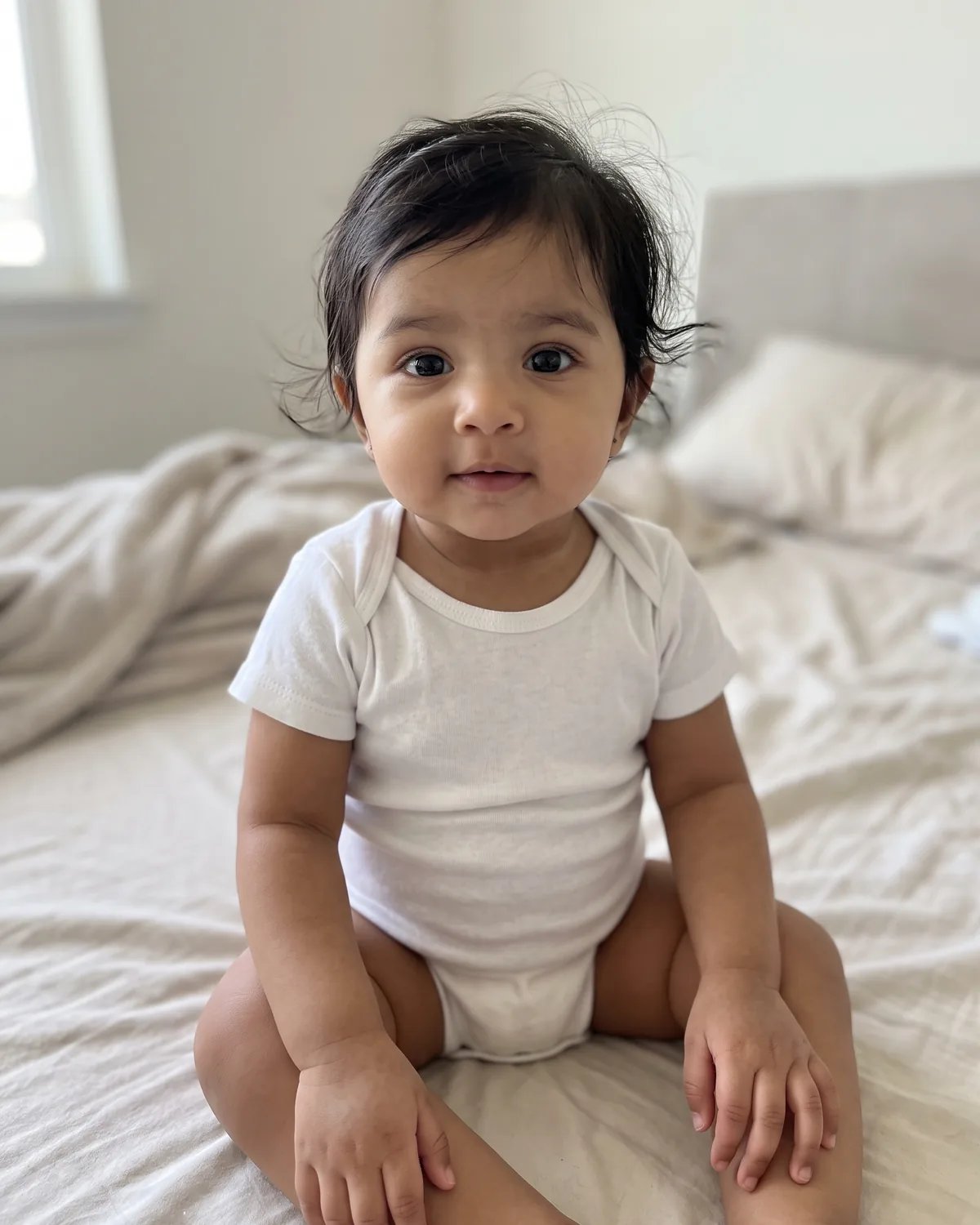 South Asian baby with wispy black hair in a white onesie, sitting on a bed with cream sheets.