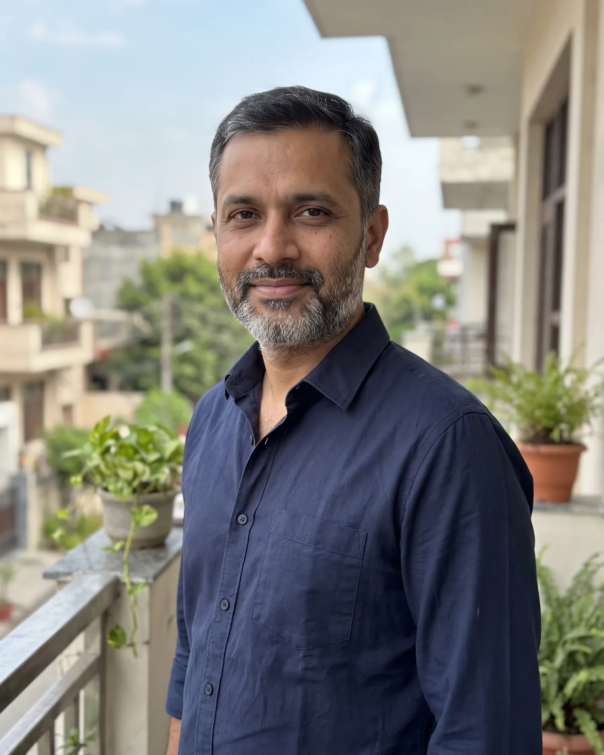 South Asian man with short hair greying at the temples and a salt-and-pepper beard in a navy button shirt, on a home balcony with plants.
