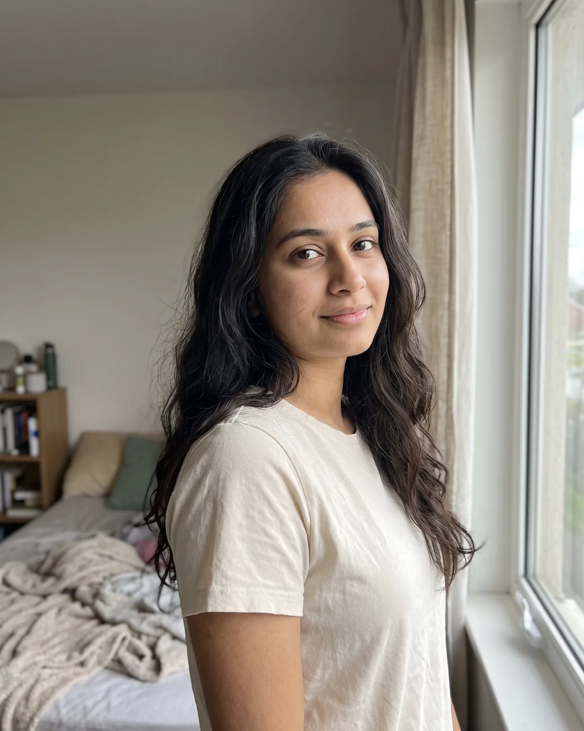 Young South Asian woman with long wavy dark hair in a cream t-shirt, standing by a bedroom window.