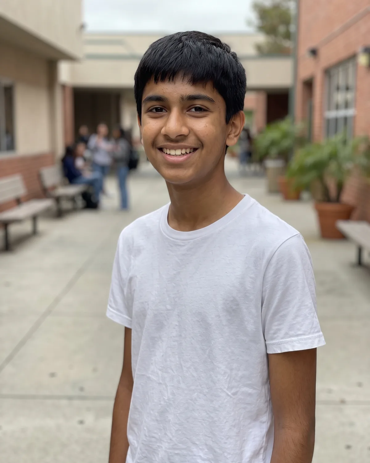 South Asian teenage boy with short straight black hair in a white t-shirt, grinning in a school courtyard.