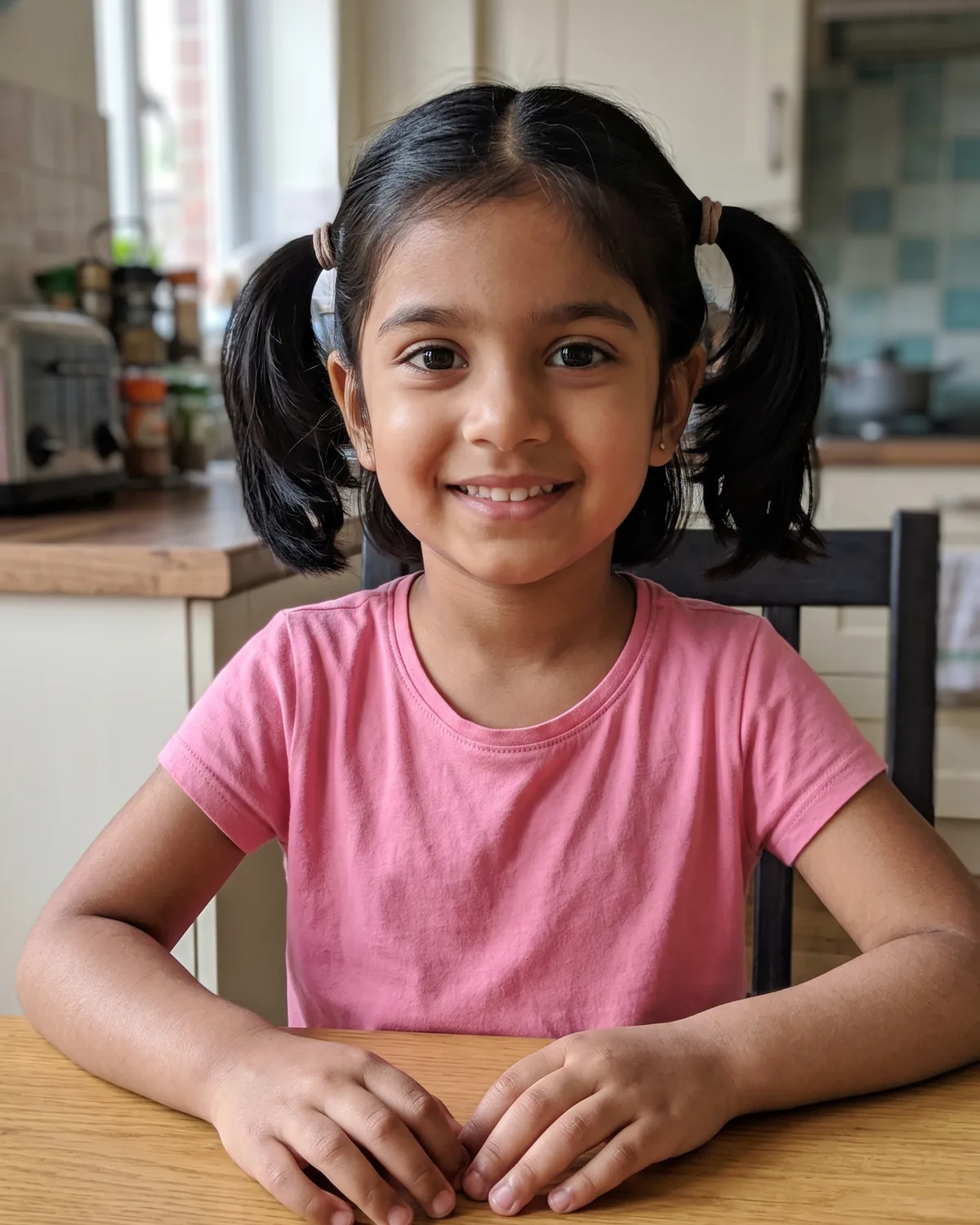 South Asian girl, about six, with black hair in pigtails and a pink t-shirt, smiling at a kitchen table.