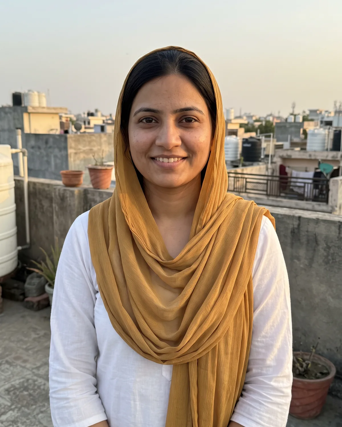 South Asian woman with a mustard dupatta over her hair and a white kurti, smiling on a city rooftop at dusk.