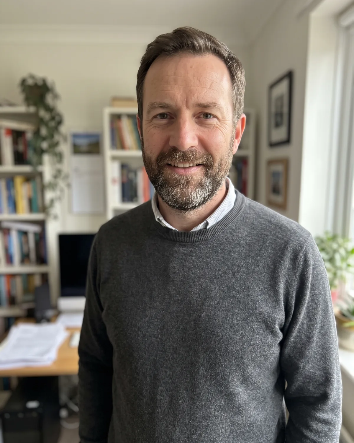White man with short brown hair and a greying beard in a charcoal sweater over a collared shirt, in a home study with bookshelves.