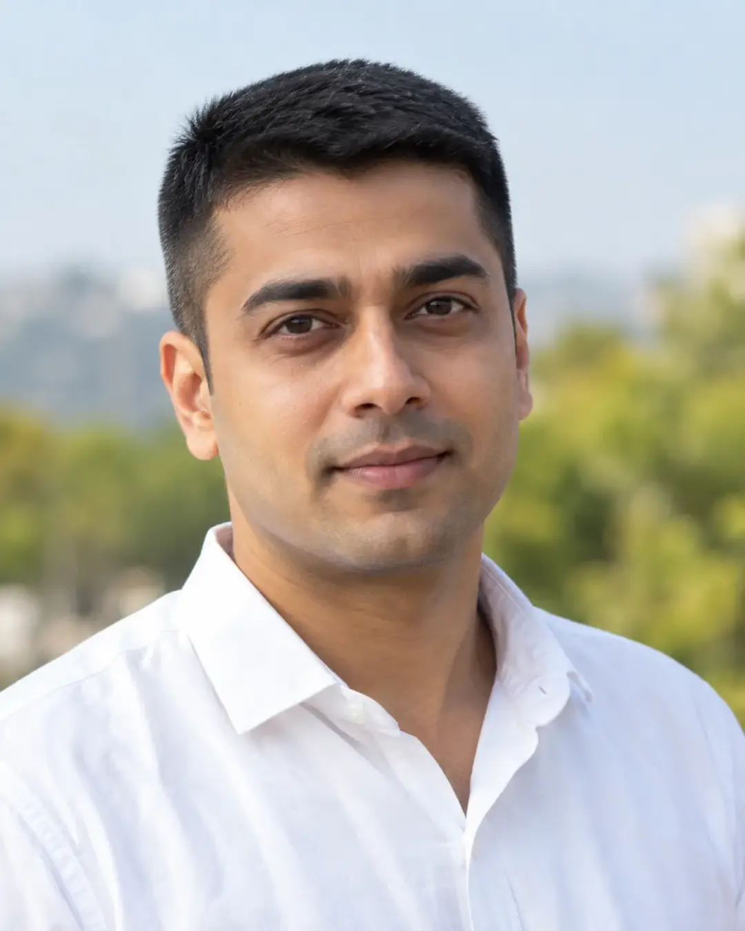 Man with short dark hair in a white open-collar shirt, outdoors with trees and a hazy skyline behind.