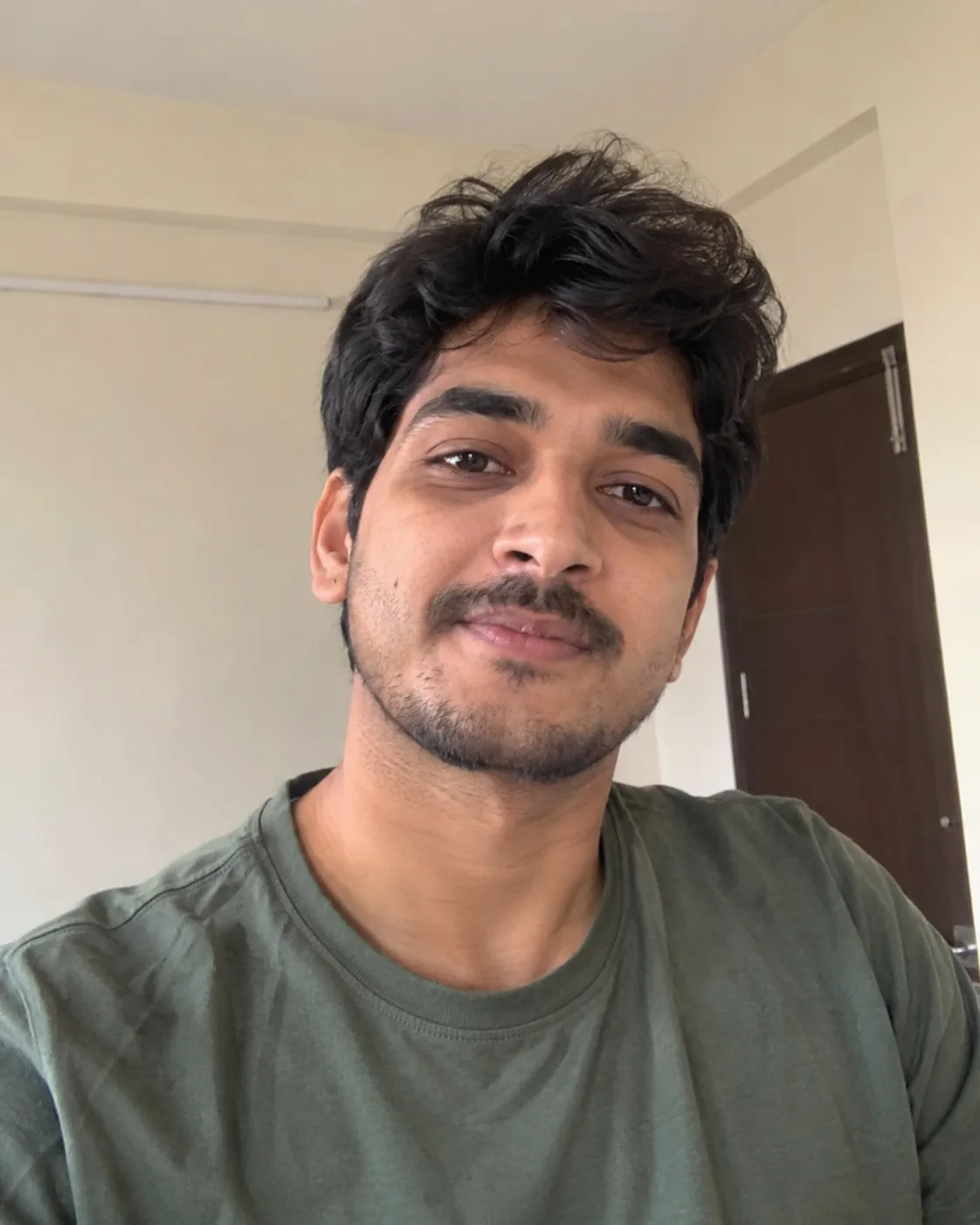 Young man with tousled dark hair and a light moustache in an olive-green tee, indoors.