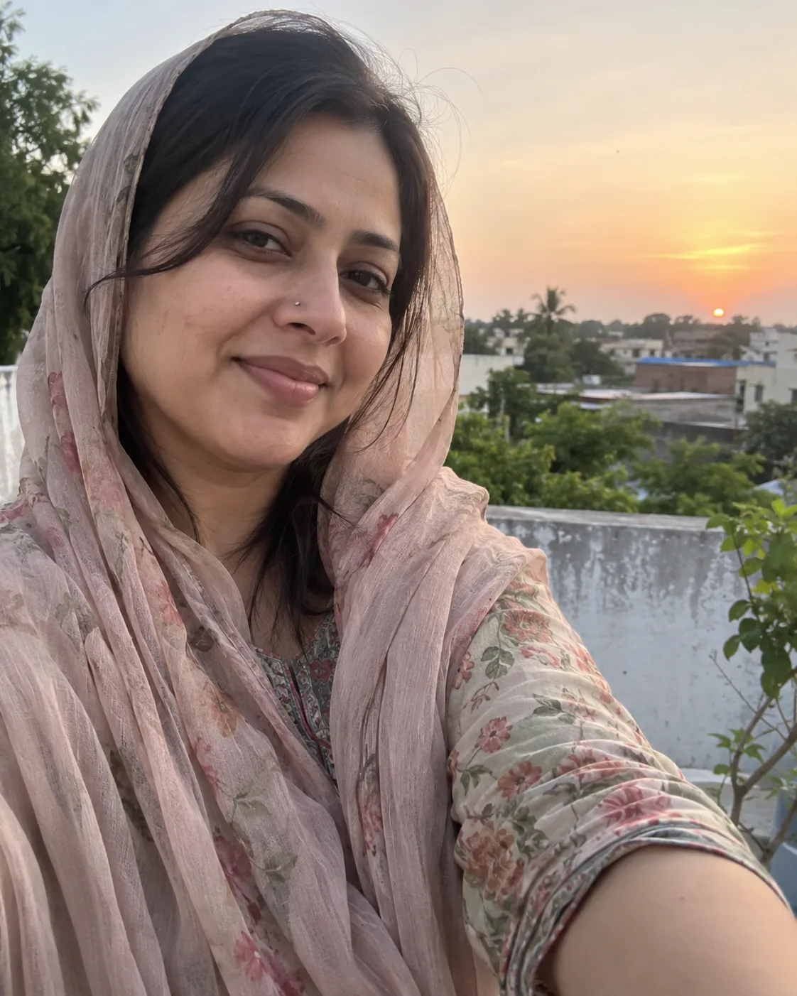 Woman in a pink dupatta and floral kurti, selfie against a sunset sky.