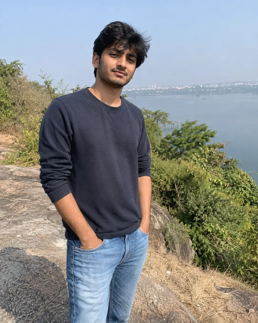 Young man in a navy sweatshirt and light jeans, with greenery and water behind.