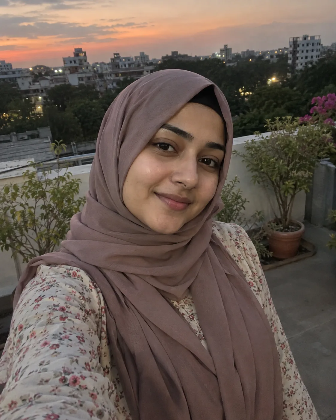 Woman in a dusty-rose hijab and floral dress, rooftop selfie at dusk.