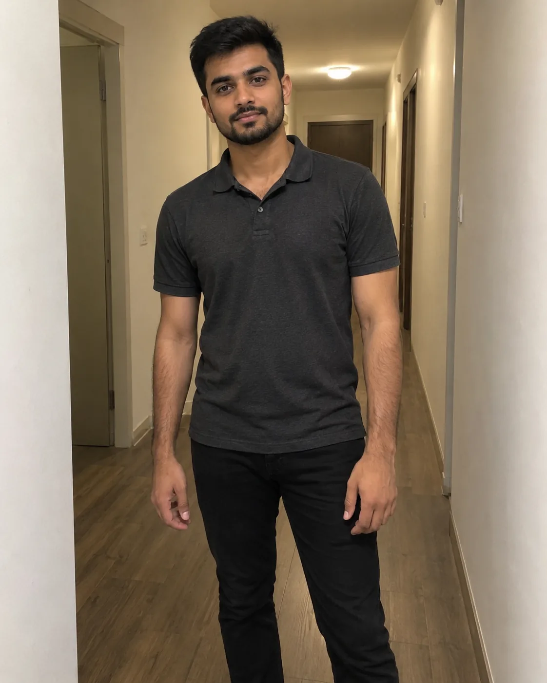Young man with a neat beard in a charcoal polo, in a beige hallway.